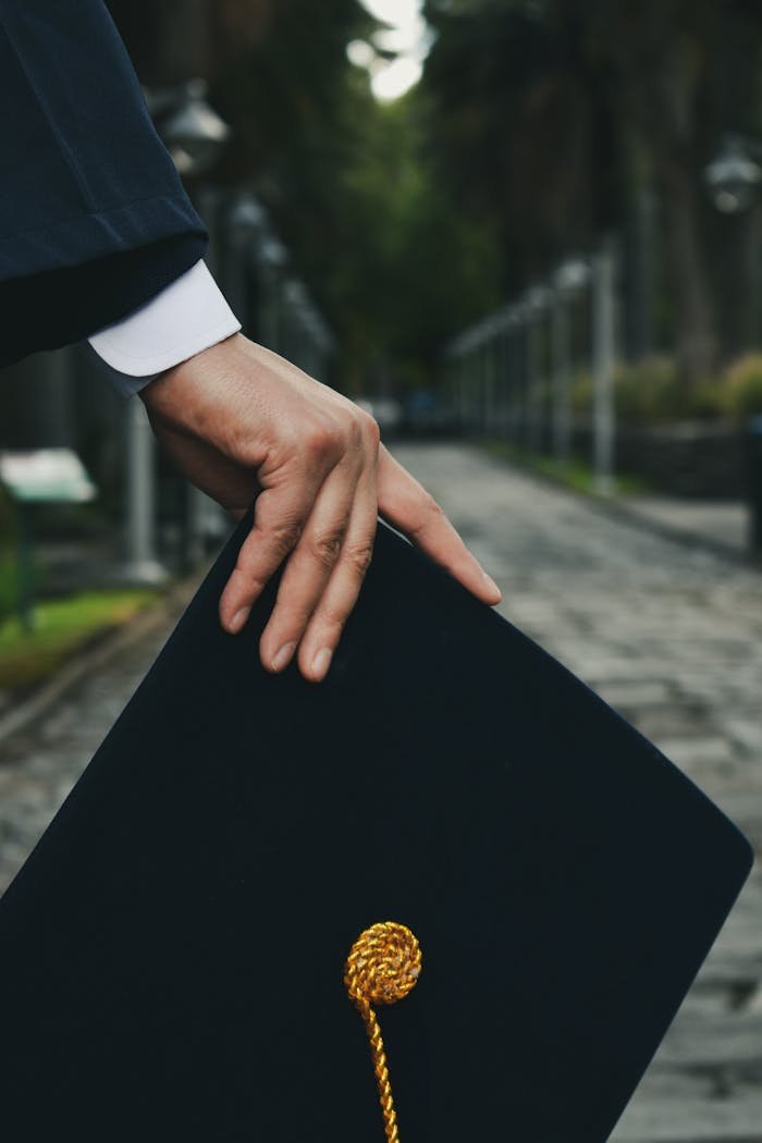 A close-up shot of a hand holding a graduation cap on an outdoor path, symbolizing achievement.