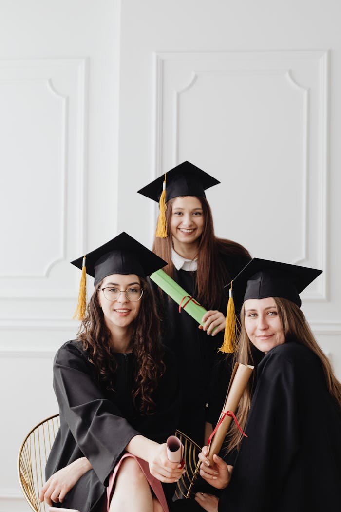 Home Three female graduates in caps and gowns posing with diplomas indoors.