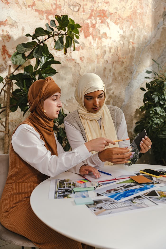 Home Two women in hijabs work together on a creative project at a table with photos indoors.